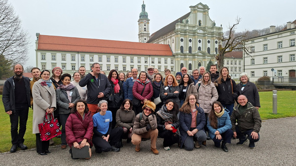 A large group of approximately 40 people posing for a group photo outdoors on a gravel path. They are wearing warm autumn/winter clothing and smiling. In the background stands a large historic building complex with a white baroque church facade and a clock tower.