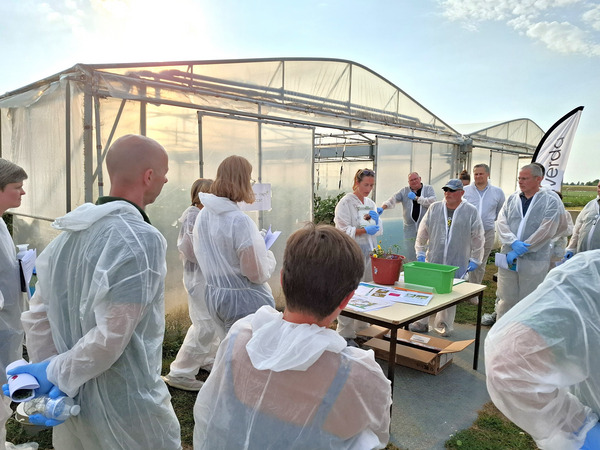 Participants wearing protective coveralls gather outside a greenhouse tunnel while a speaker demonstrates cultivation practices using plants and materials displayed on a table.