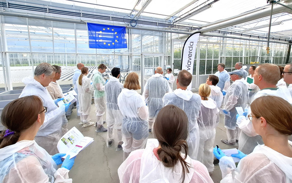 A group of participants wearing white protective coveralls stands inside a greenhouse during a guided tour. An EU flag and a Viaverda banner are visible above the group.
