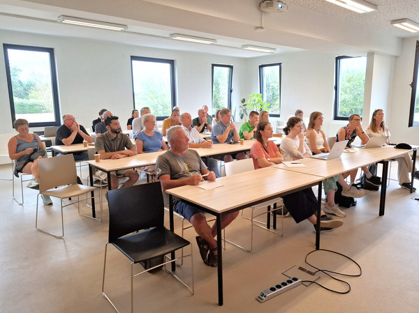 Participants seated at tables in a meeting room attend a presentation during the field trial visit. Several attendees take notes or work on laptops while facing the speaker.